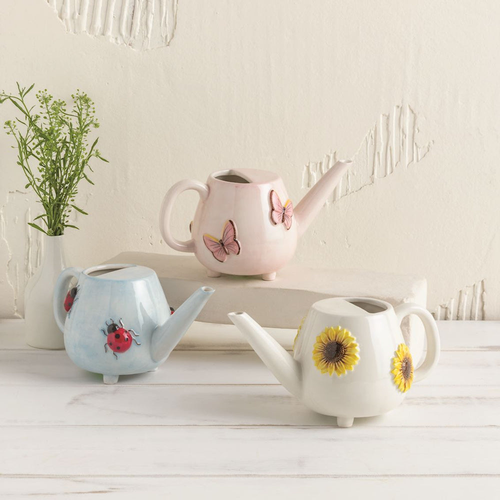Three ceramic watering cans in pastel pink, blue, and white, decorated with 3D butterflies, ladybugs, and sunflowers.