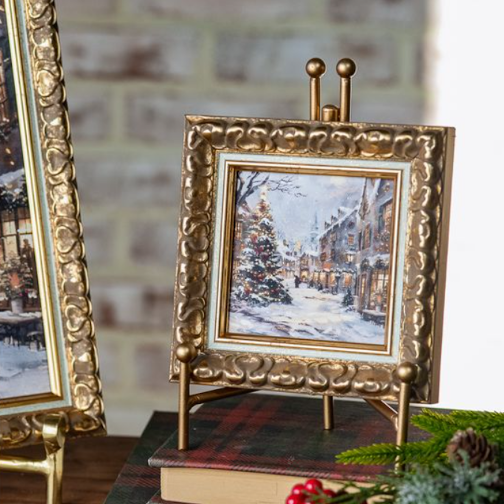 Small framed artwork of a snowy Christmas village scene displayed in an ornate gold frame on a stand.