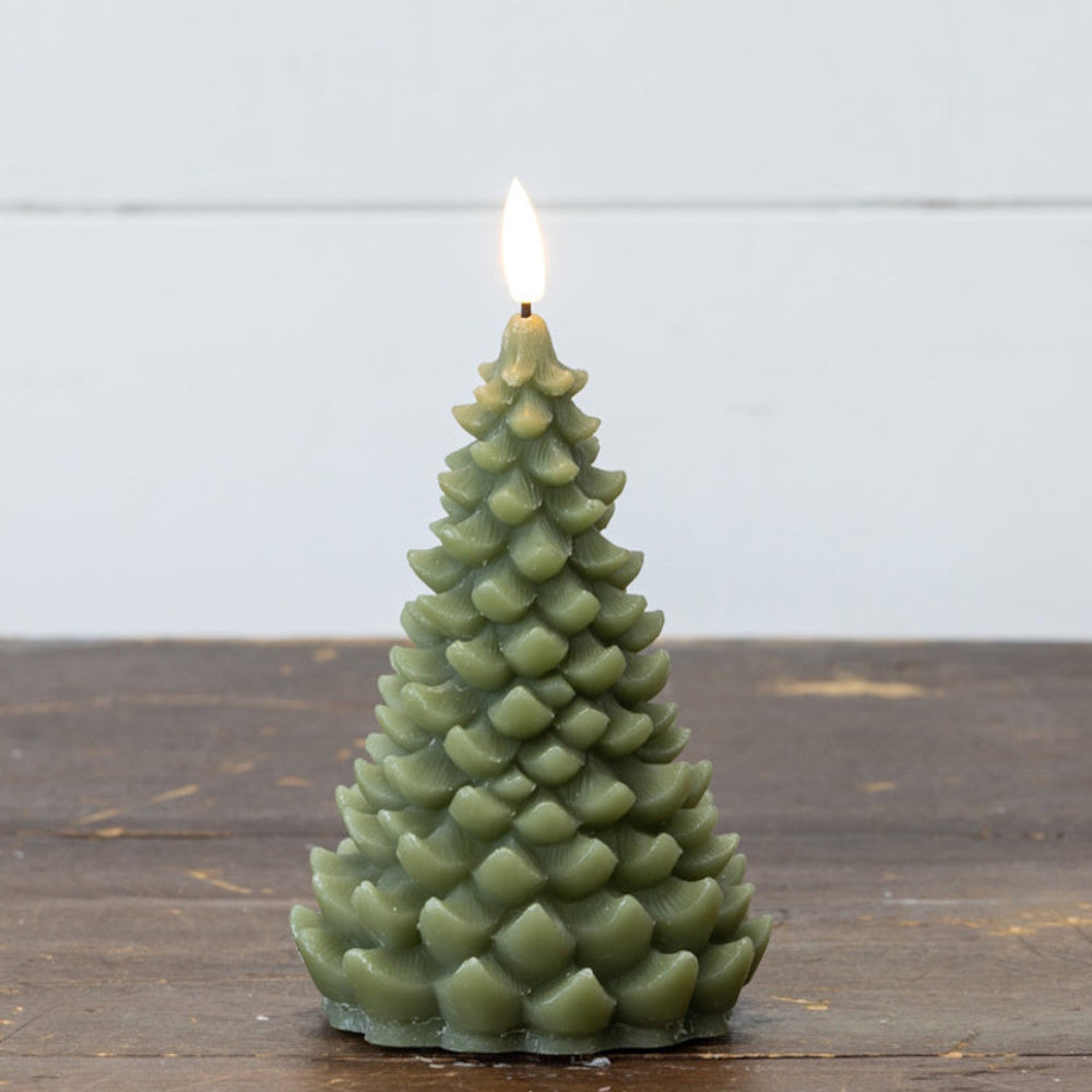 A green, pine tree-shaped candle with a lit flame, sitting on a rustic wooden table against a white background.
