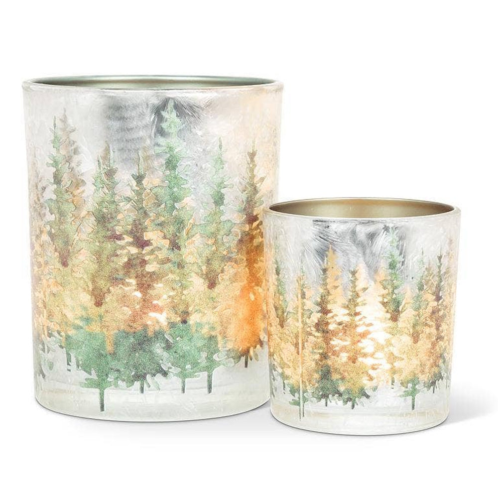 Two frosted glass candle holders featuring glowing, glittery winter forest pine tree scenes.