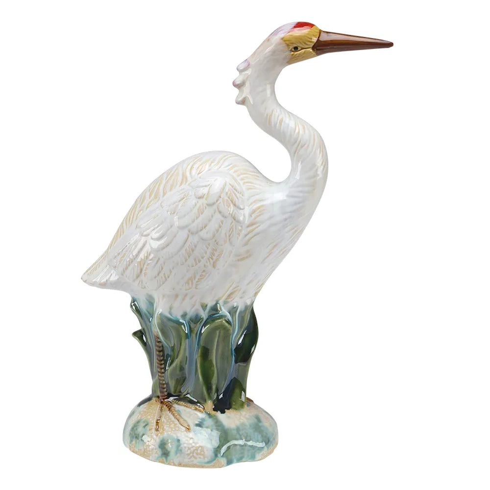 Glossy ceramic heron figurine with white feathers and a red crown, standing in green marsh grass.