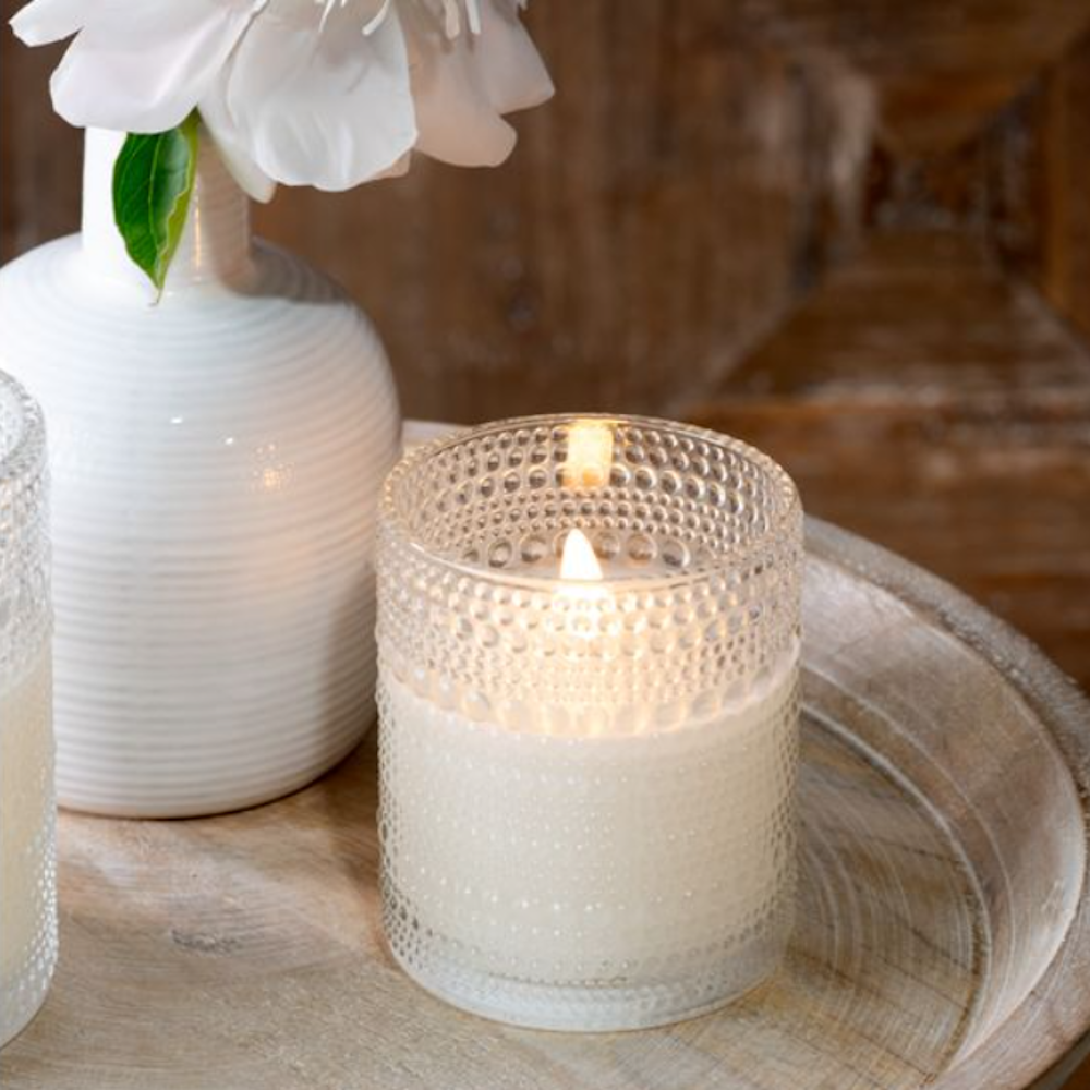 Lit LED candle inside a clear, hobnail beaded glass holder on a wooden tray next to a vase.