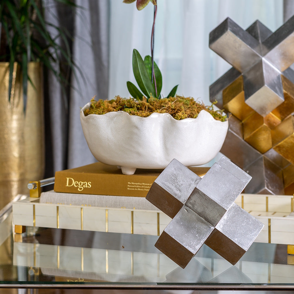 Modern white wavy ceramic bowl with moss and orchid on a stack of books and a decorative silver cross sculpture.