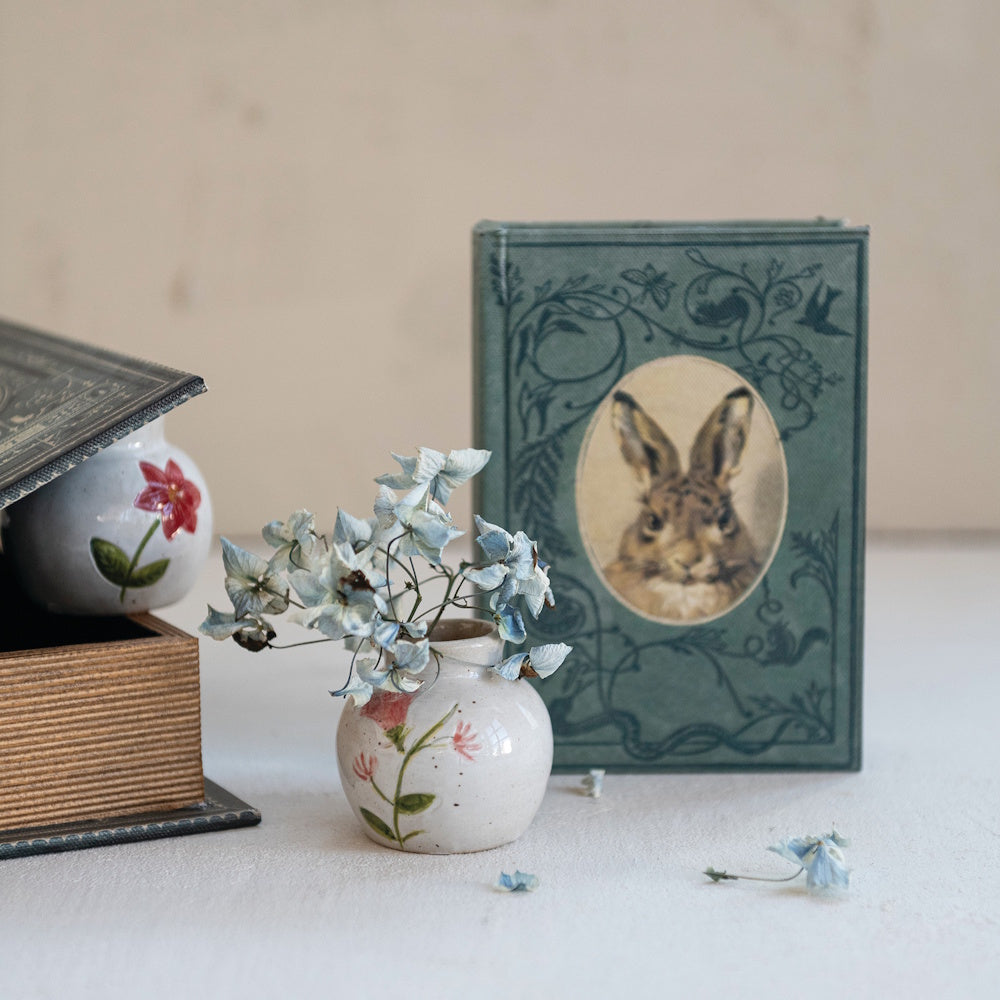 A vintage book featuring a rabbit illustration alongside a small ceramic vase holding blue dried flowers.