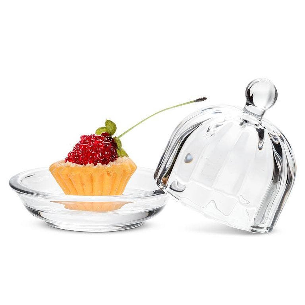 A small clear glass dish with an optic ribbed lid next to a miniature fruit tart topped with a red berry.