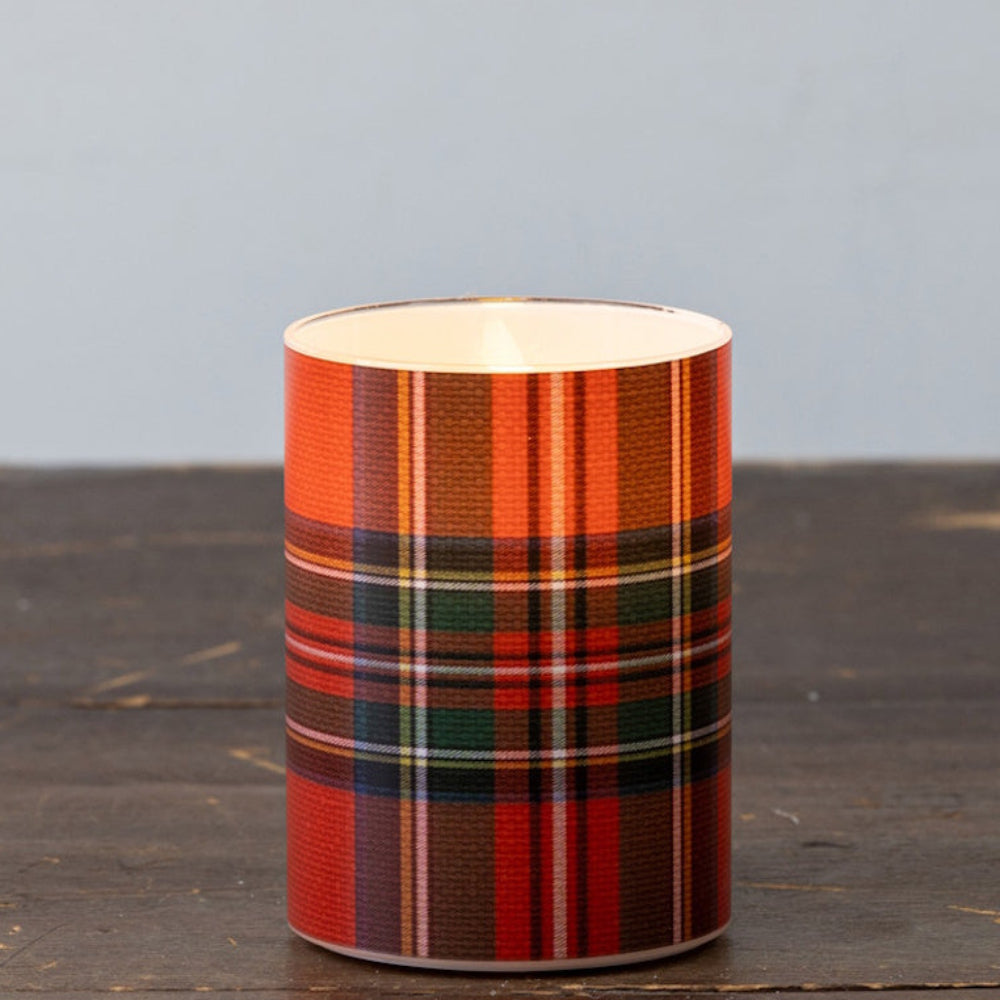 A lit glass candle wrapped in a red plaid tartan pattern resting on a rustic wooden surface.
