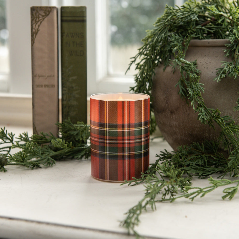 Lit red tartan plaid glass candle on a windowsill next to vintage books and pine greenery.