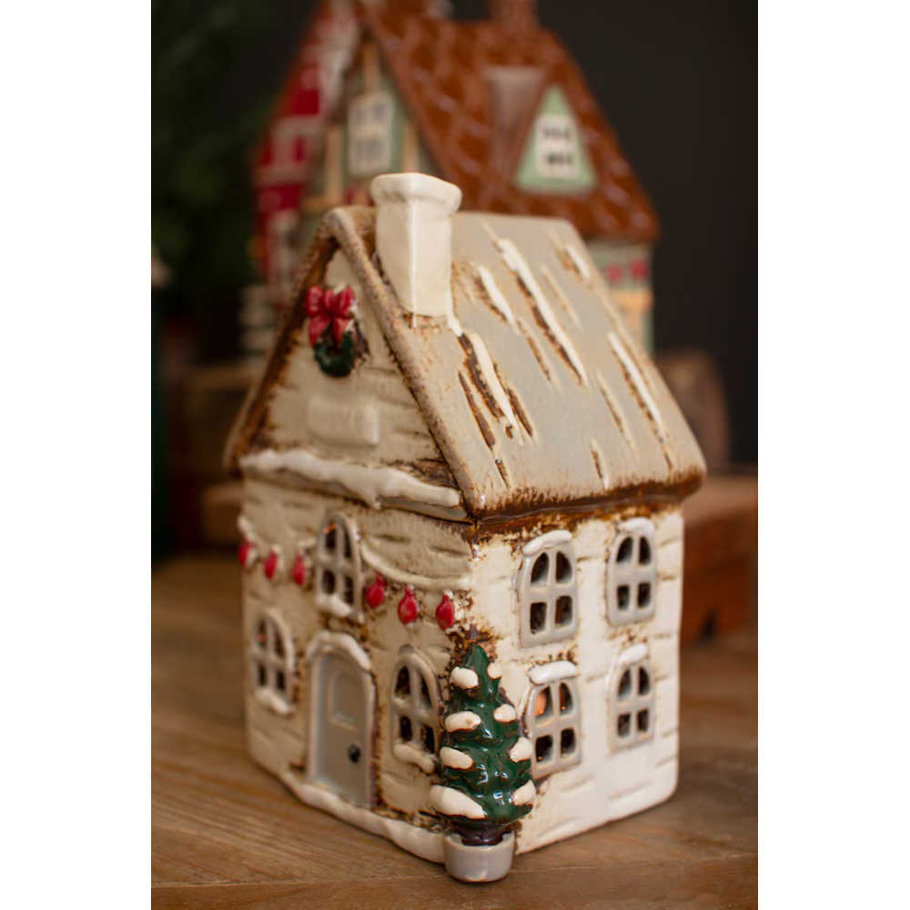 Side-angle view of a cream ceramic Christmas village house with snow-dusted roof and tree details.
