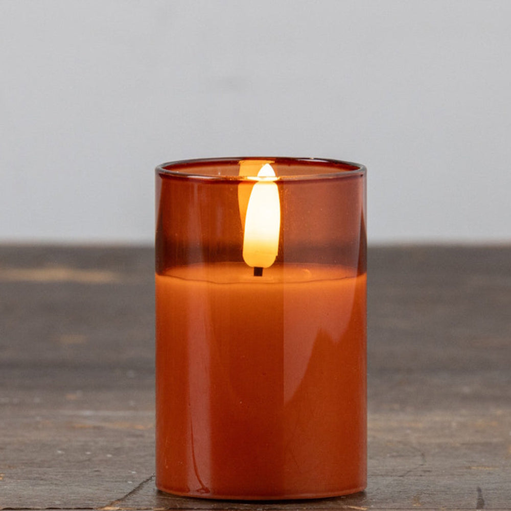 A glowing flameless LED candle inside a small sienna glass holder resting on a rustic wooden table.