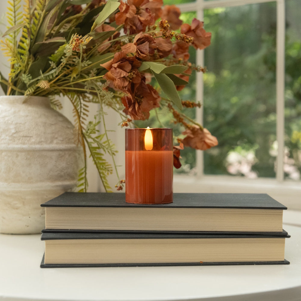 Sienna glass flameless candle set on stacked books next to autumn floral decor near a window.