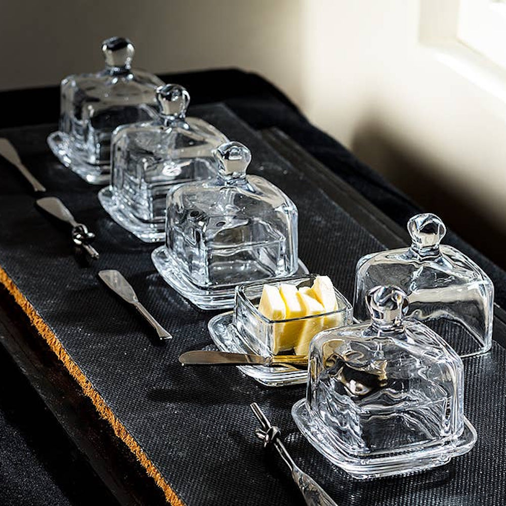 Clear glass mini covered butter dishes lined up with small spreader knives on a dark tray.