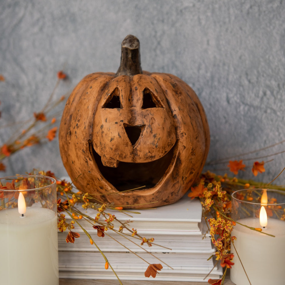 A carved terracotta jack-o'-lantern on stacked books surrounded by candles and autumnal floral accents.