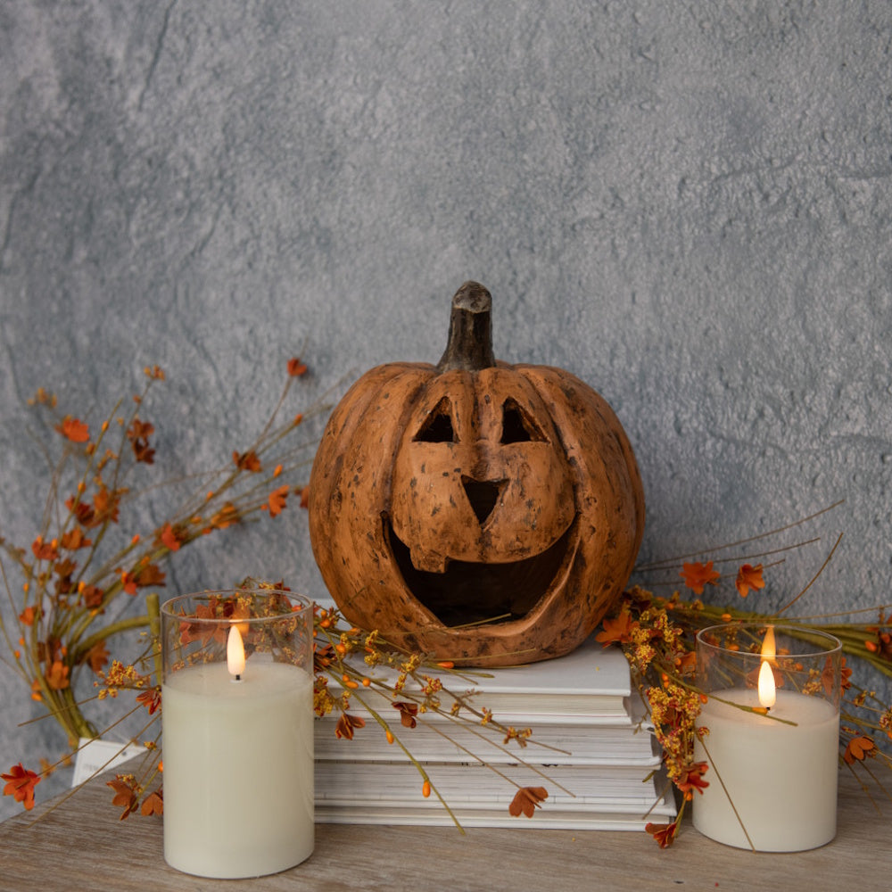 Terracotta jack-o'-lantern on stacked books flanked by two glowing candles and autumn florals.