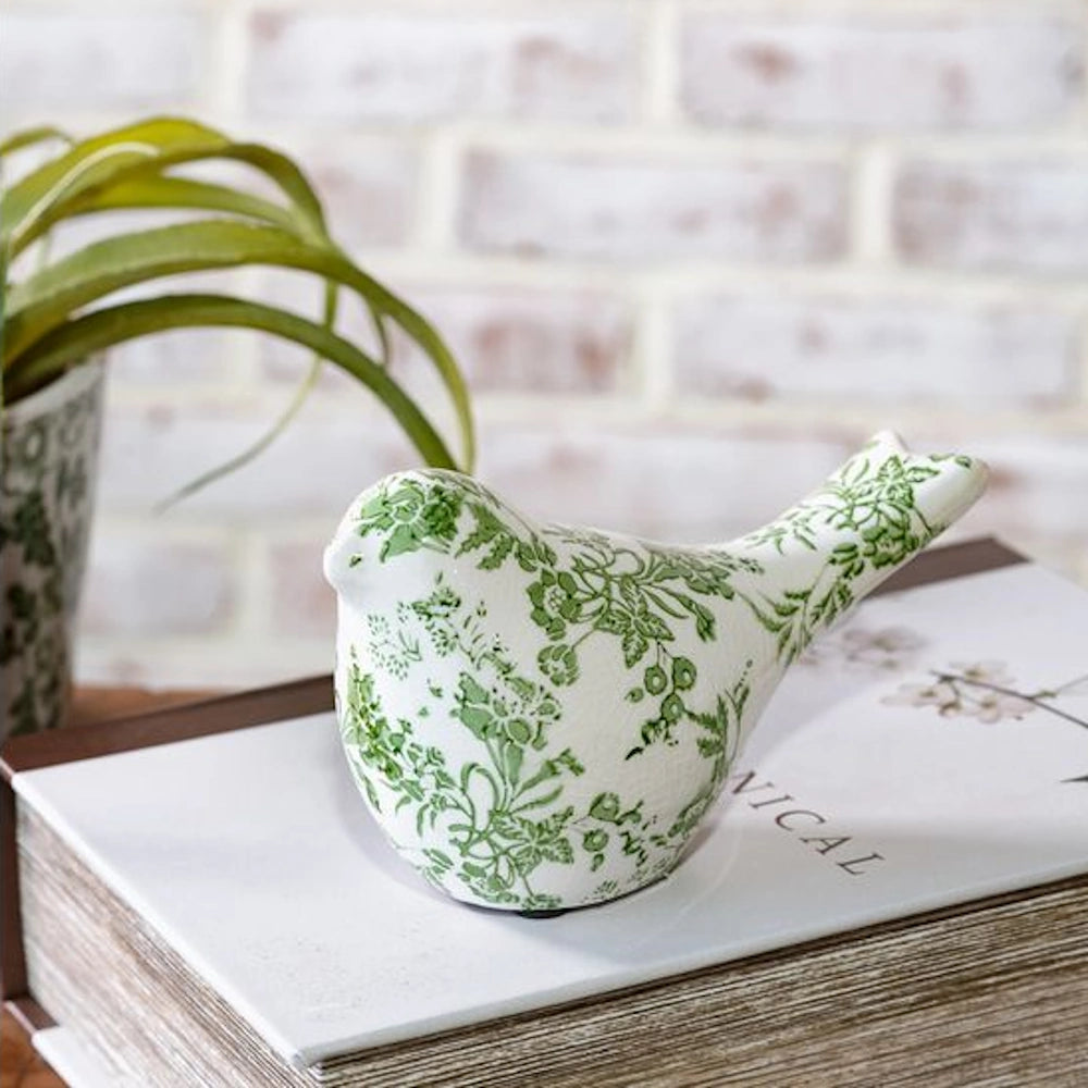 Ceramic bird figurine with a green floral toile pattern resting on a white book against a blurred brick background.