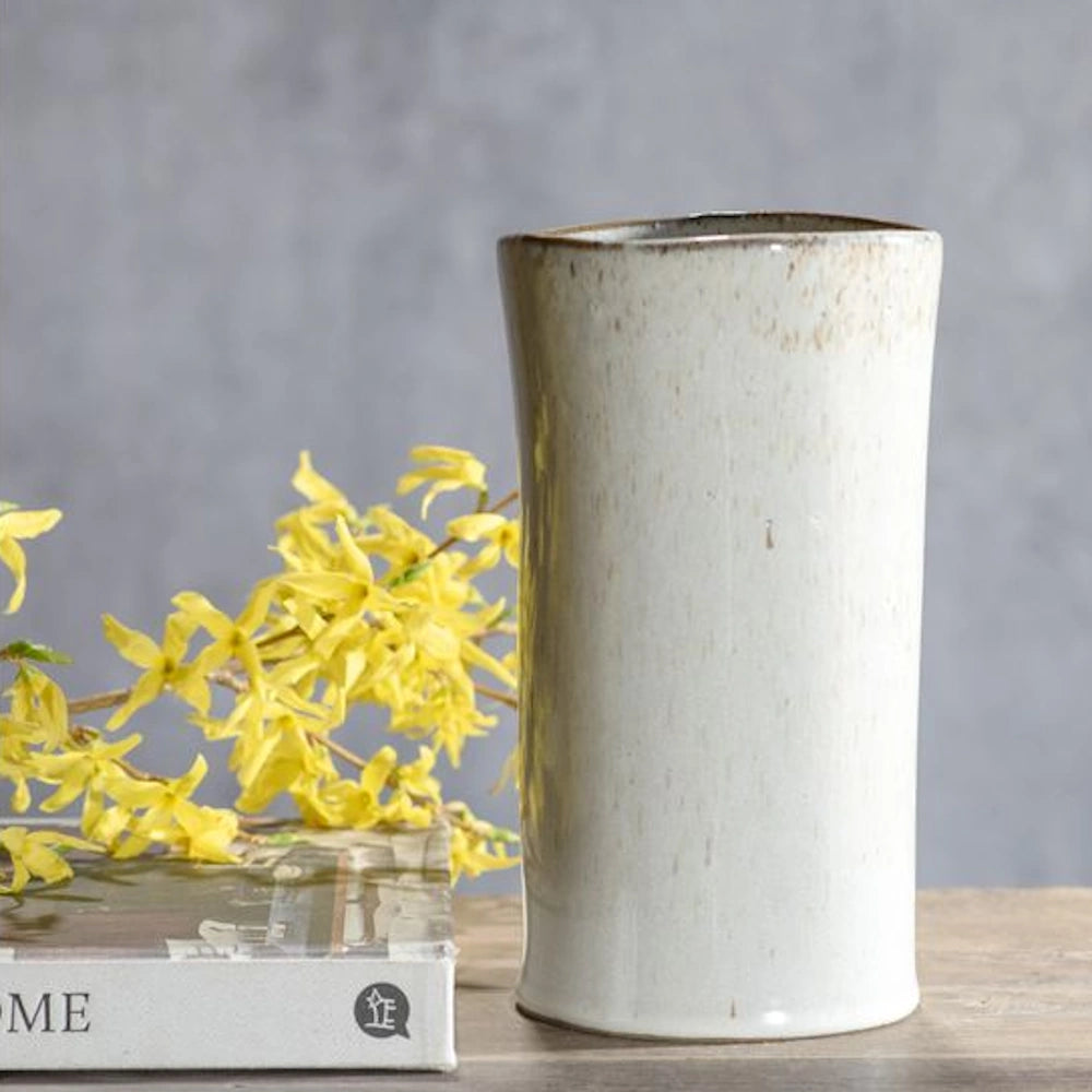 White sand ceramic vase with speckled glaze standing next to yellow forsythia flowers on a wooden table.