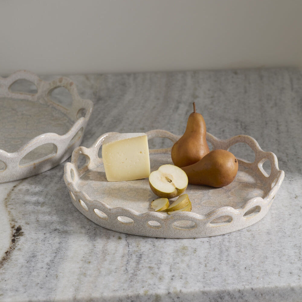 A ceramic tray with a scalloped cutout rim holds cheese and Bosc pears on a marble countertop.