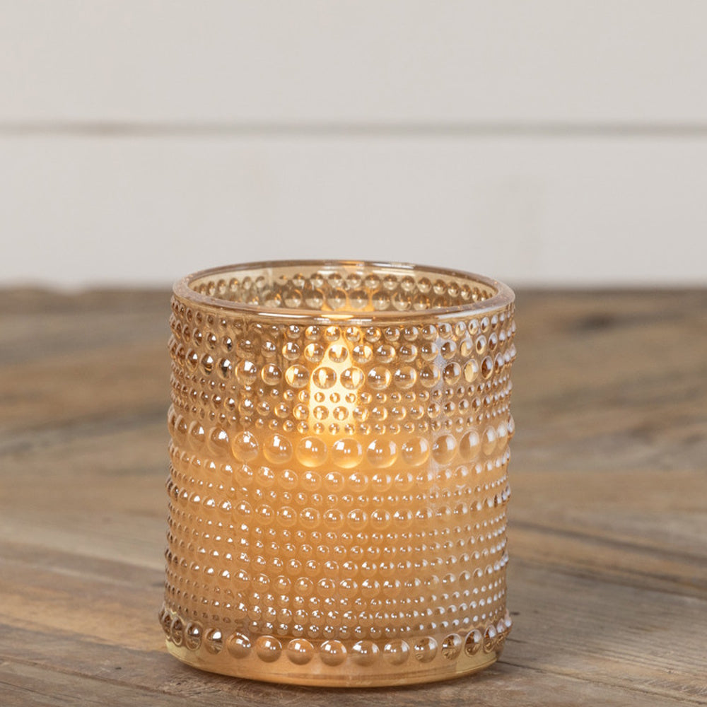 Lit candle inside a champagne-gold beaded glass holder resting on a wooden surface.