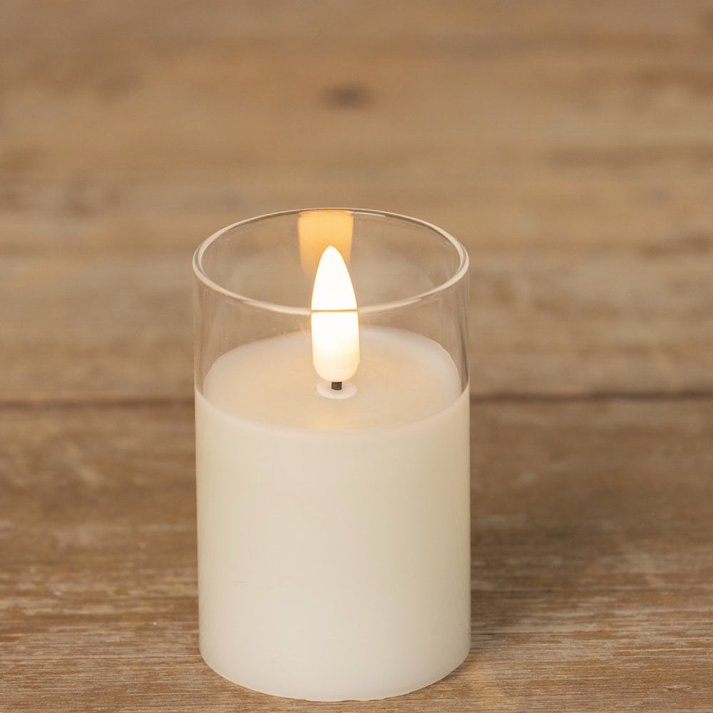 Lit flameless white candle inside a clear glass cylinder, set on a wooden surface.