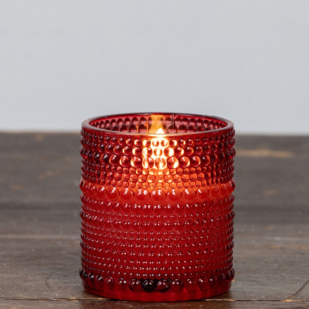 Small red hobnail glass candle holder with a glowing flame inside, sitting on a wooden surface.