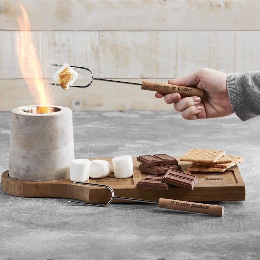 A hand roasting a marshmallow over a mini concrete tabletop fire pit next to a s'mores ingredient board.