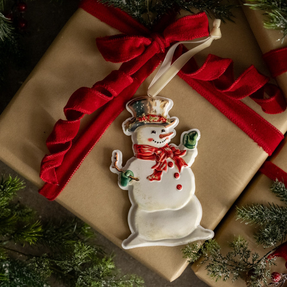 A snowman ornament atop a brown wrapped present tied with a wavy red ribbon, framed by pine branches.