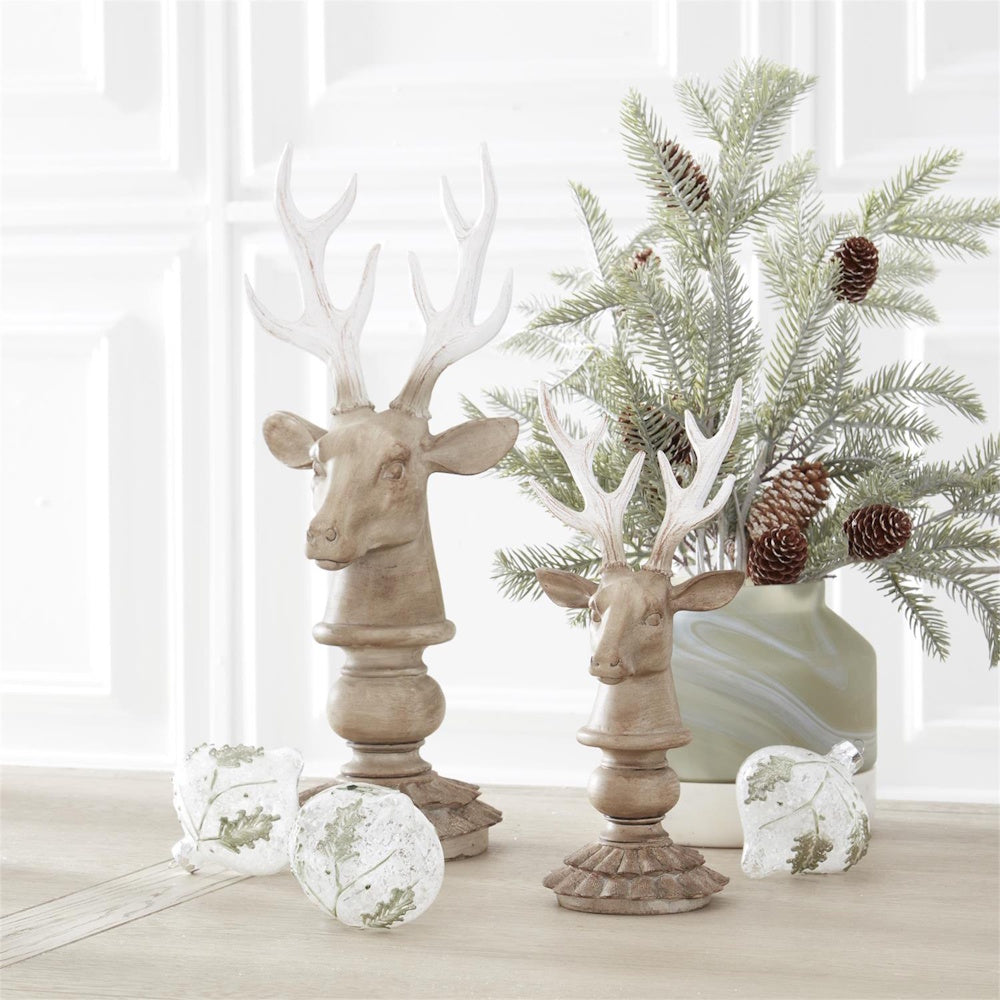 Two deer bust figurines with white-tipped antlers on stands, snowy pine branches with pinecones, and clear glass ornaments.