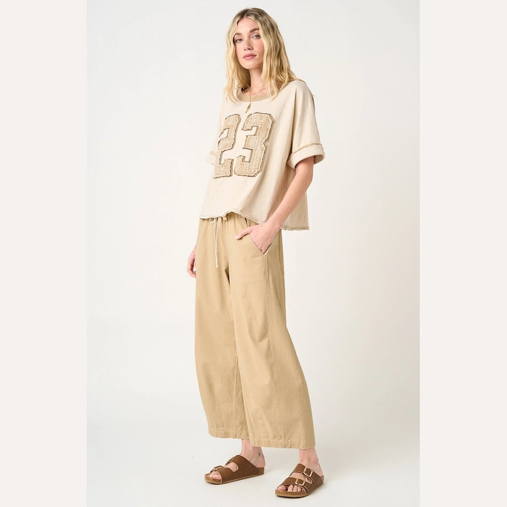 Woman wearing tan wide-barrel drawstring pants, a beige graphic tee with the number 23, and brown slide sandals.