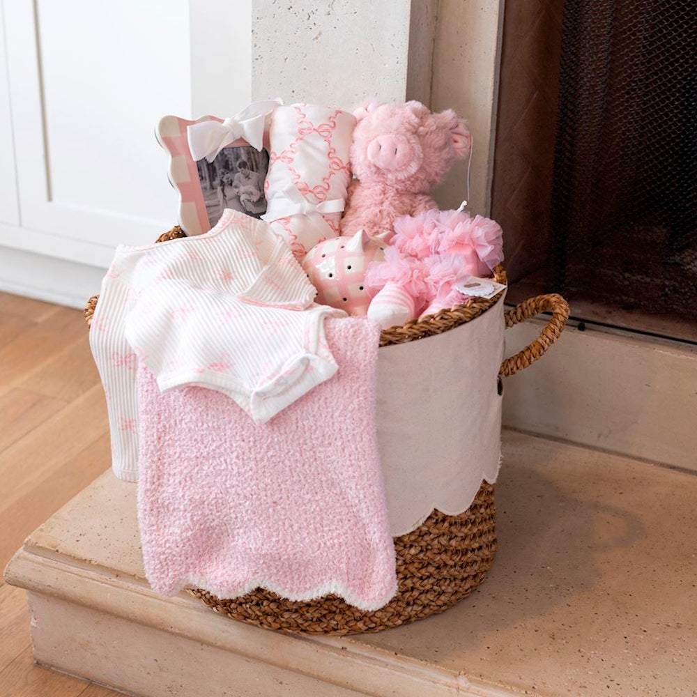 Pink baby gift basket with swaddle, plush pig, and scalloped blanket by a fireplace.