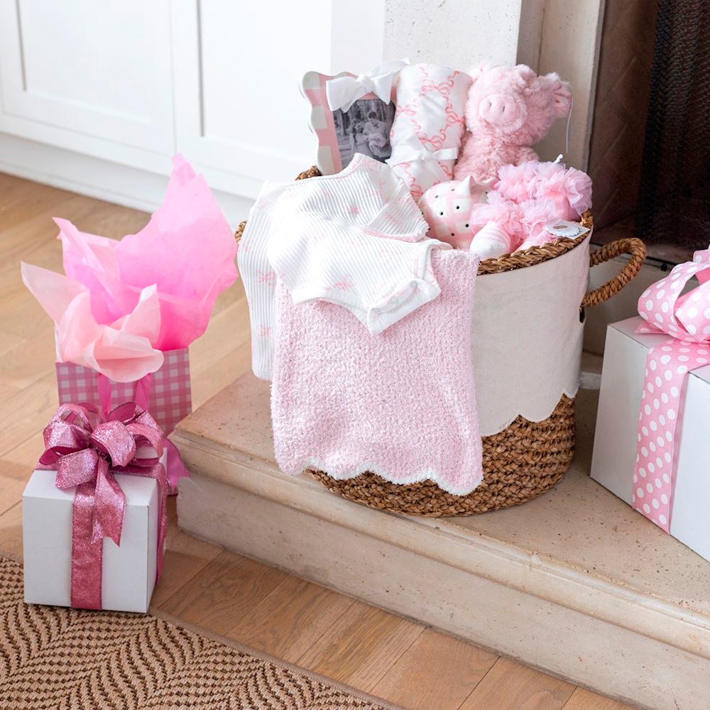 Pink baby gift basket with a soft knit swaddle, plush pig toy, and scalloped blanket next to wrapped gifts.