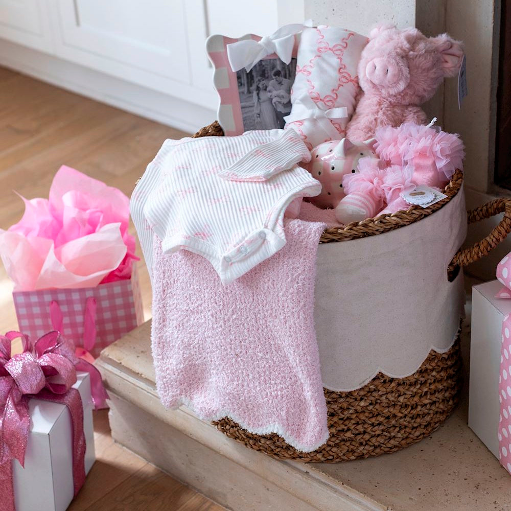 Pink baby girl gift basket with plush pig, striped onesie, and soft knit blankets in a woven scalloped bin.