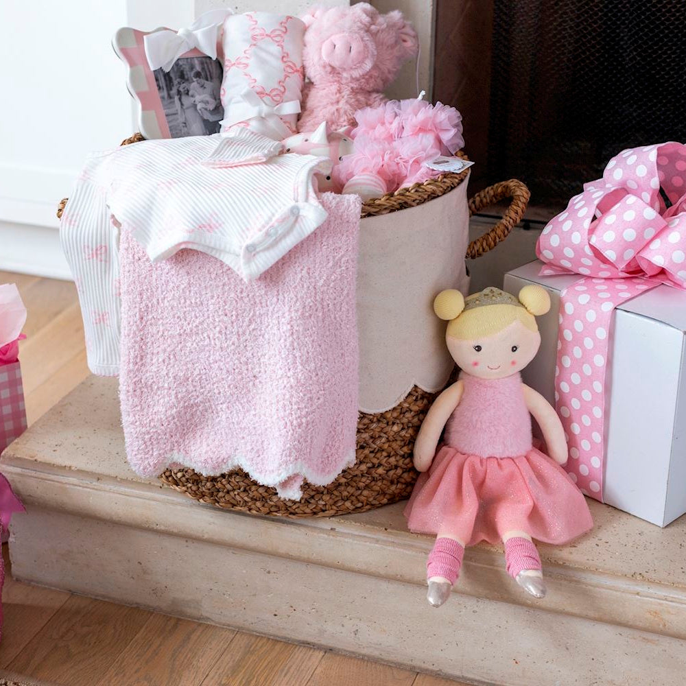 Pink baby girl gift basket with plush toys, a ballerina doll, soft blankets, and clothes in a woven scalloped hamper.