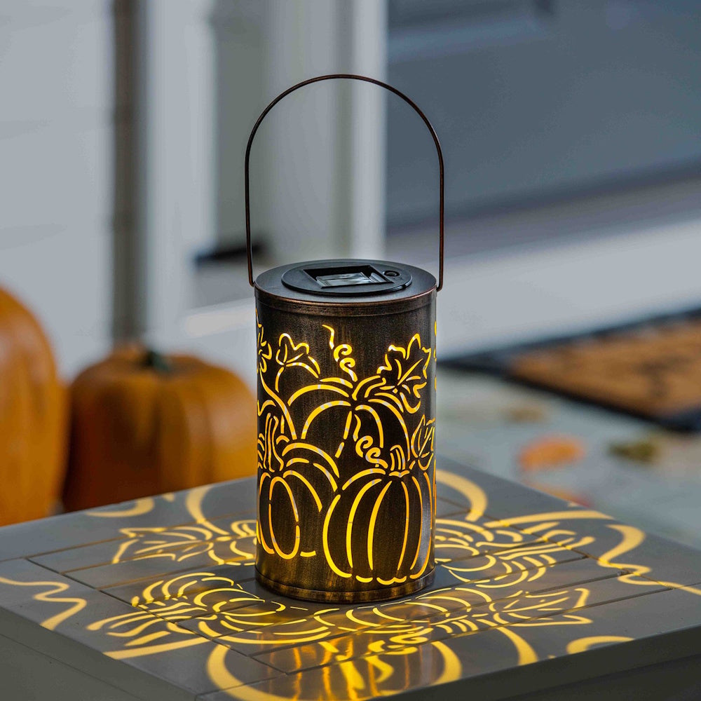 A bronze, solar-powered lantern with pumpkin cutouts glows from within, casting warm, patterned light.