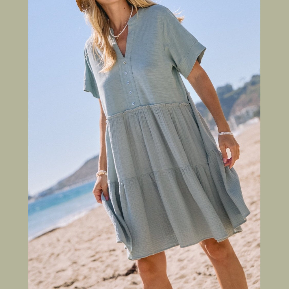 Woman wearing a light blue tiered mini dress with short sleeves and buttons, standing on a sandy beach.