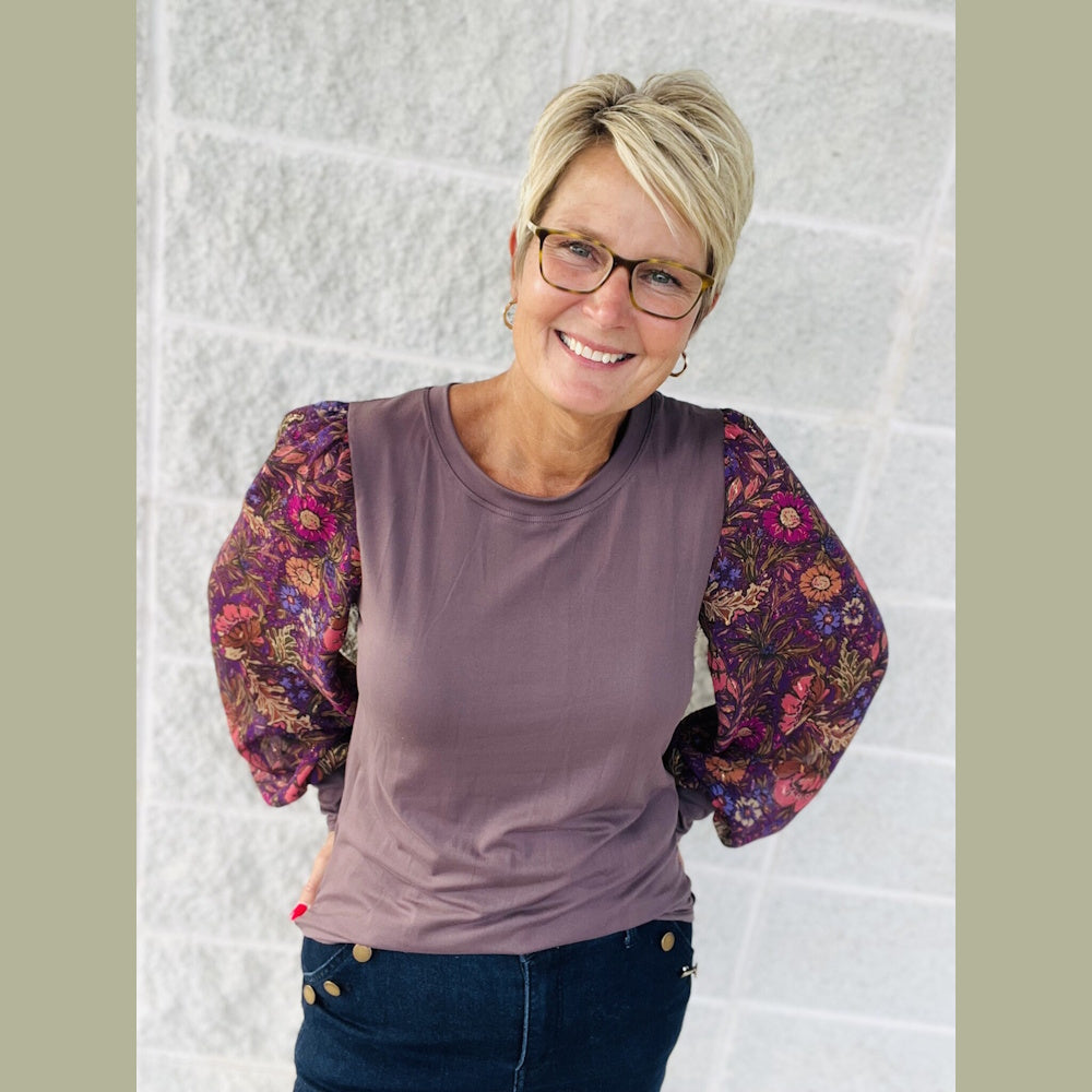 A smiling woman with short blonde hair and glasses wearing a purple top with puff floral sleeves.
