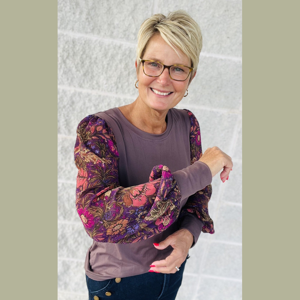 Smiling woman wearing a mauve top with colorful, balloon-style floral print sleeves and glasses.