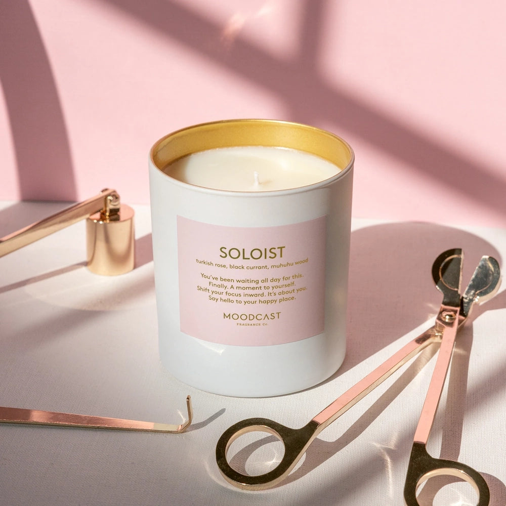 Moodcast Soloist candle with a pink label, surrounded by gold candle care tools on a sunlit surface.