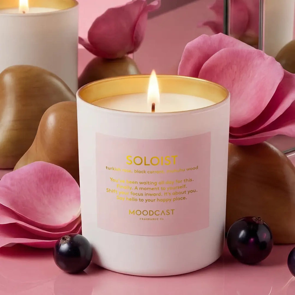 Lit Moodcast Soloist candle surrounded by pink rose petals, black currants, and wooden accents.