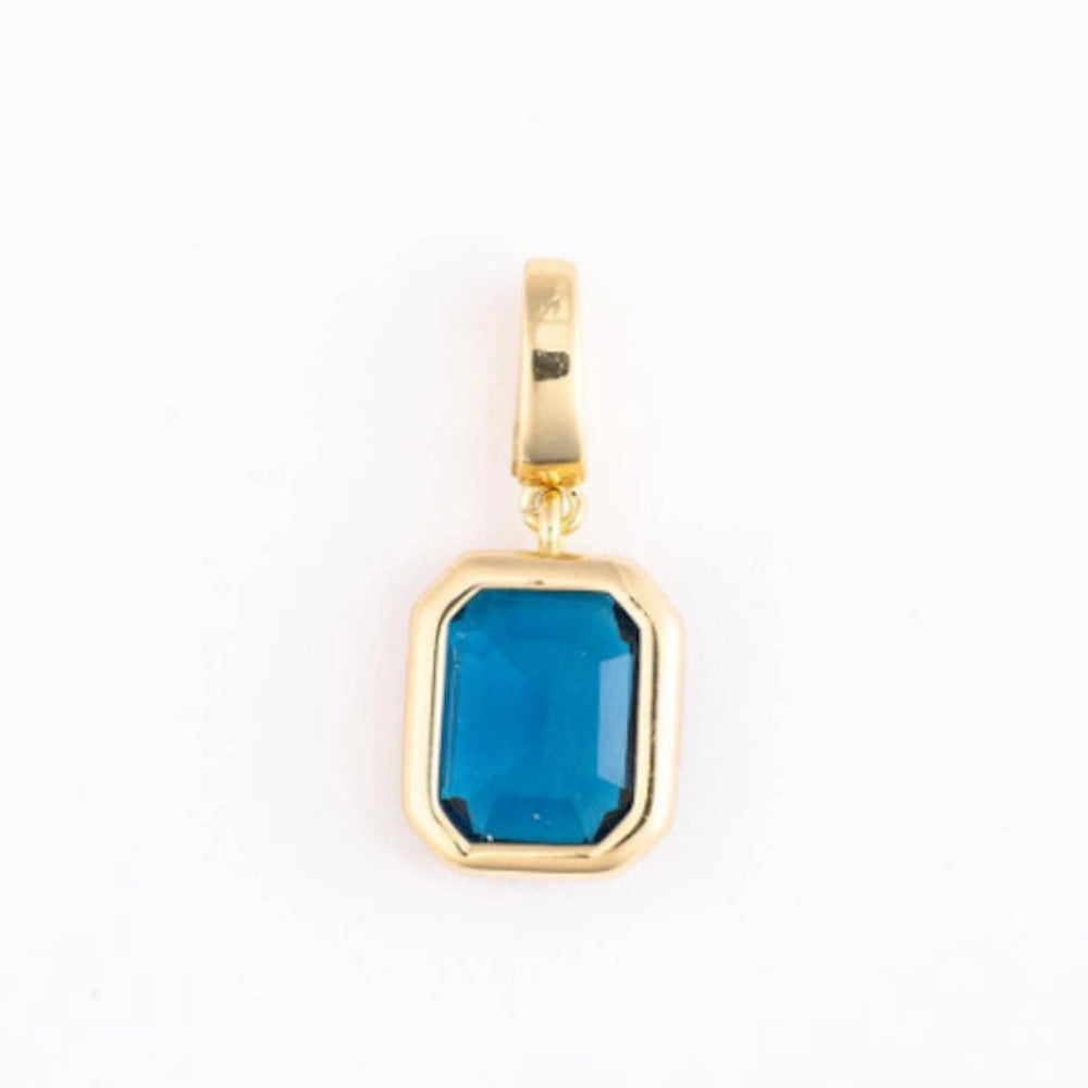 A polished gold pendant charm with an emerald-cut blue gemstone on a plain white background.