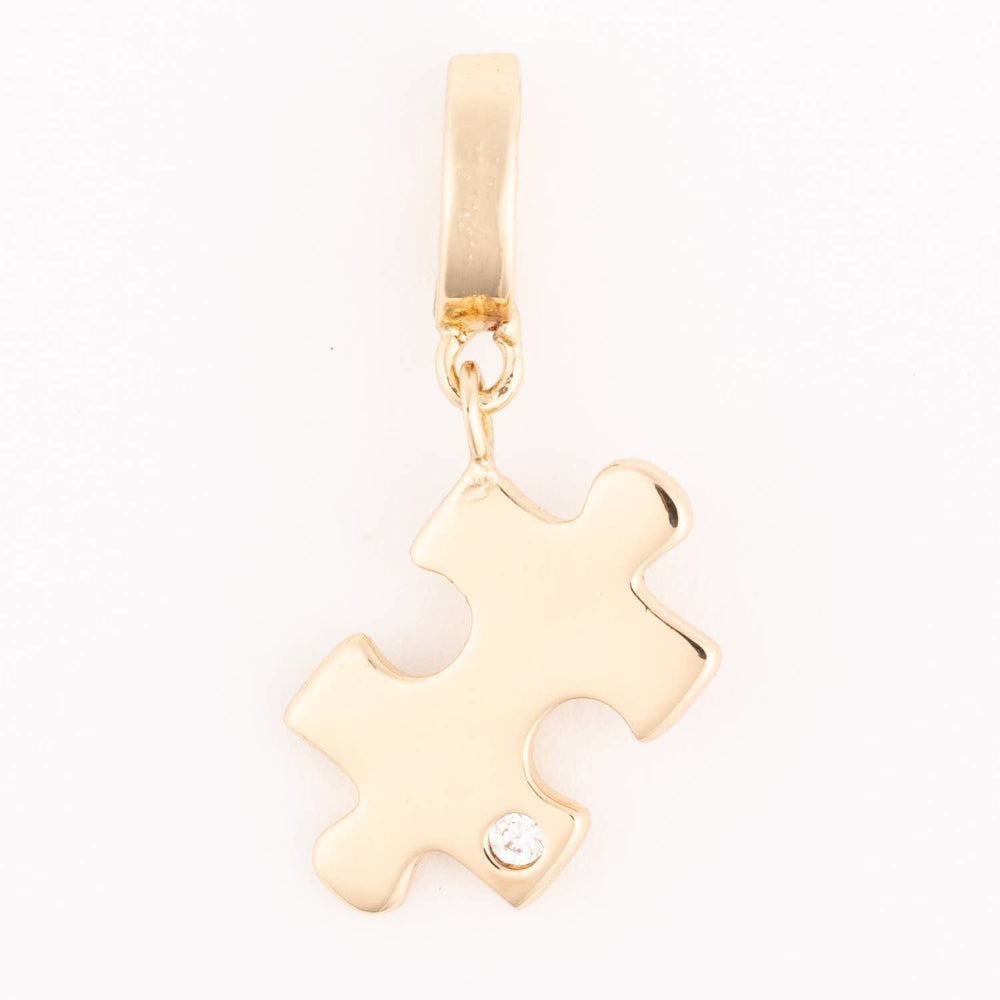 A polished gold pendant charm in the shape of a puzzle piece with a small clear stone near the bottom corner on a plain white background.