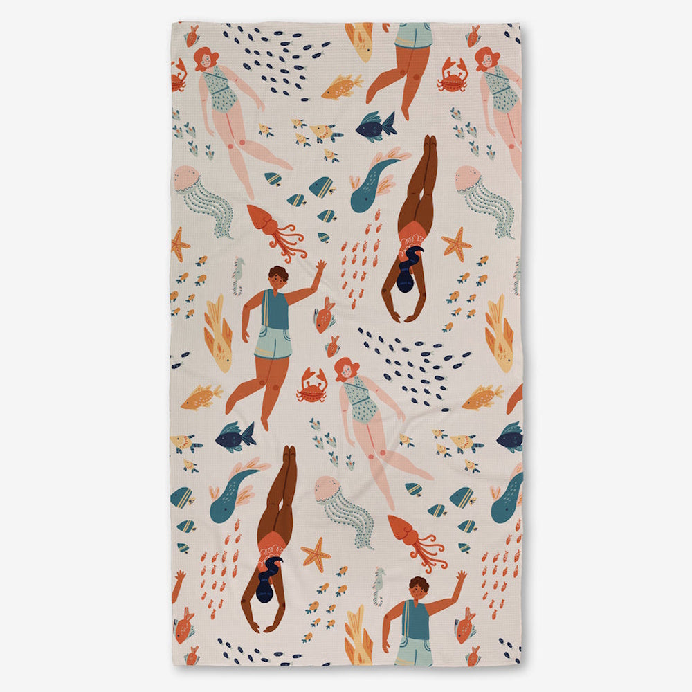 A patterned bath towel featuring an illustration of swimmers, fish, jellyfish, and starfish on a light background.