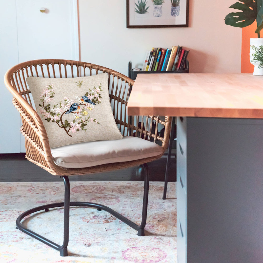 Woven chair with a floral bird-themed pillow sitting next to a wooden desk in a bright, modern room.