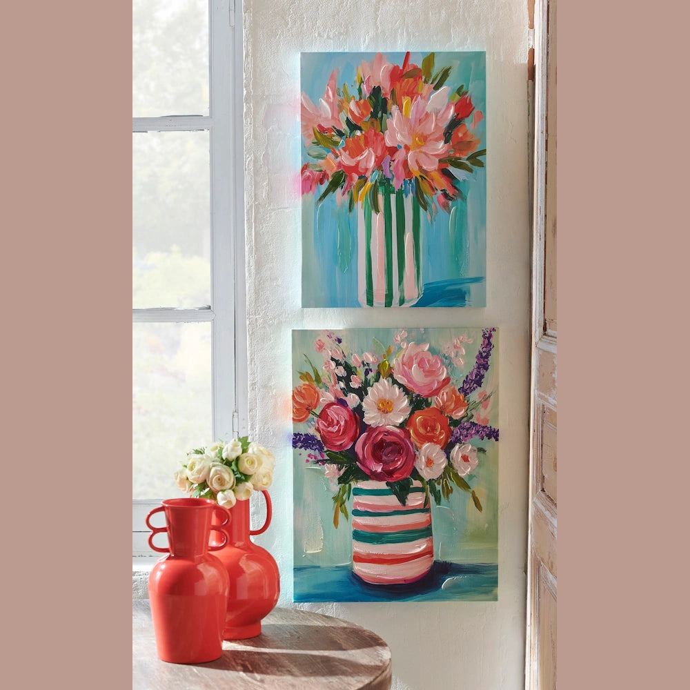 Two colorful impressionist floral paintings on a white wall above two red vases and a bouquet of white roses.