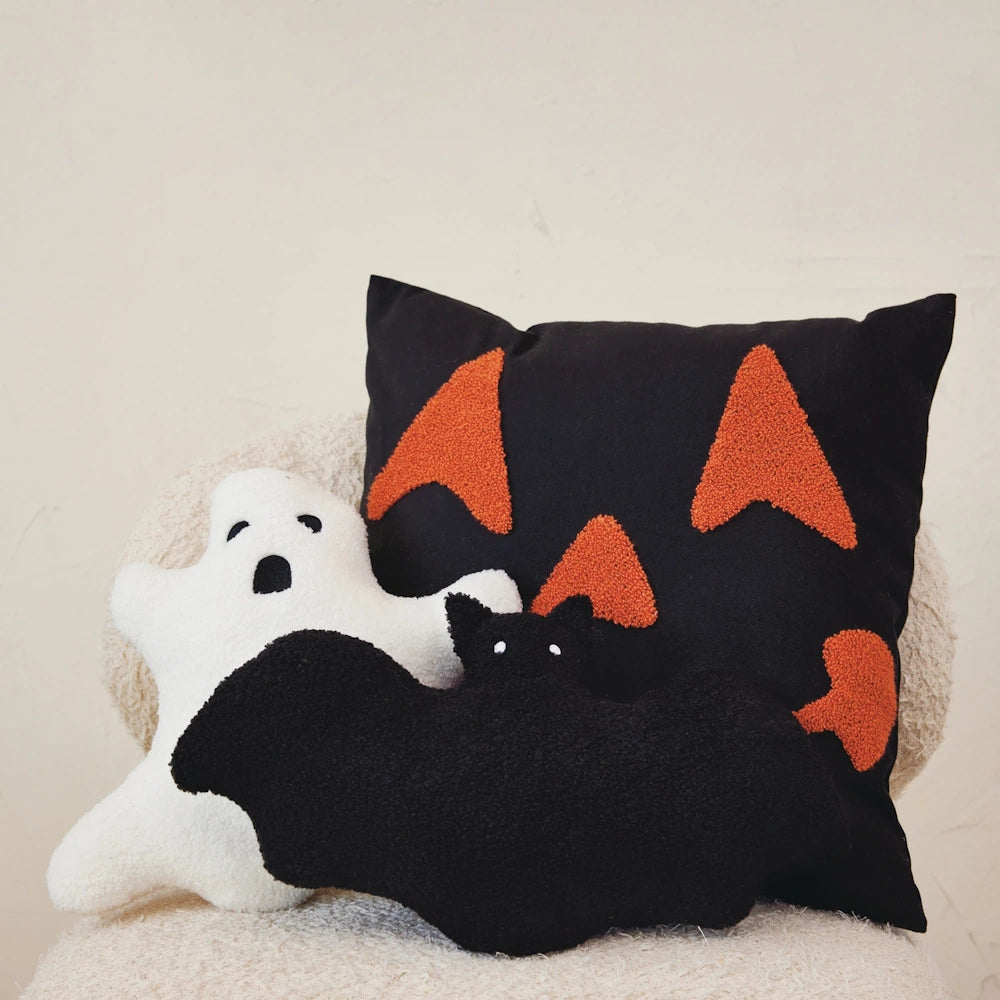 Halloween throw pillows featuring a black jack-o'-lantern, a white ghost, and a black bat on a chair.