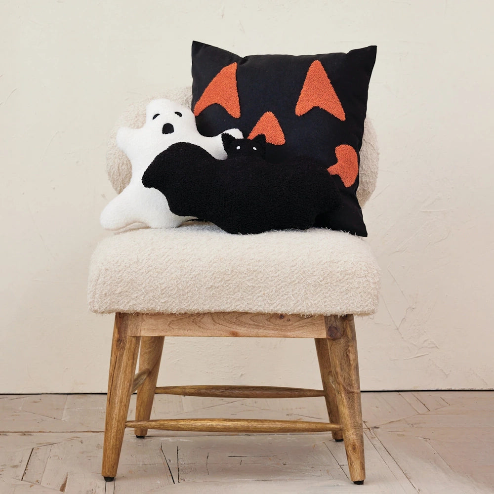 Halloween pillows including a black jack-o'-lantern, a white ghost, and a black bat on a cream boucle chair.