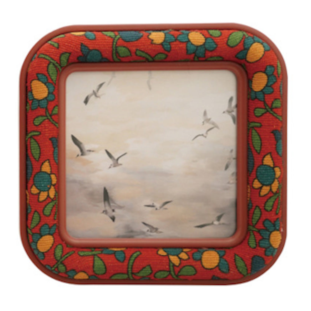Square frame with embroidered orange and green flowers, containing a picture of birds flying in a cloudy sky.