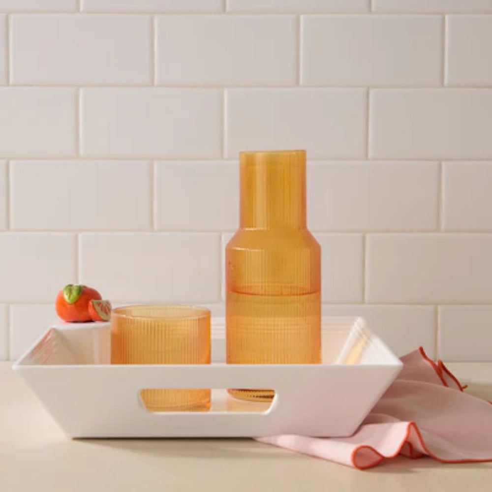White melamine tray with handles holding an amber ribbed glass carafe and tumbler against a white subway tile backsplash.