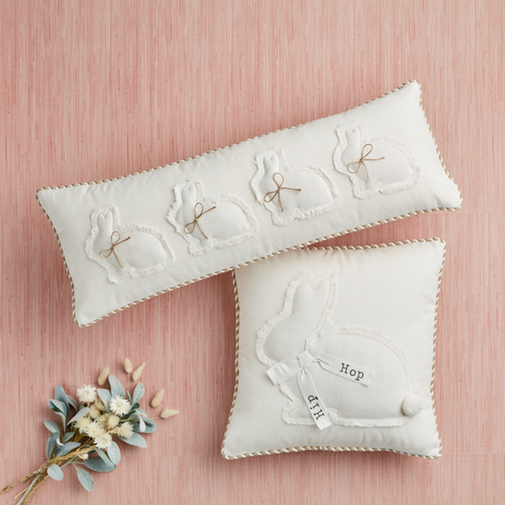 Two cream pillows with rabbit appliques and a small bouquet against a pink background.