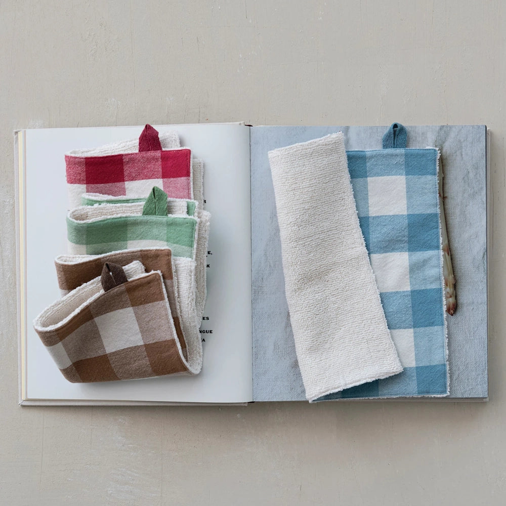 Folded gingham dish cloths stacked on an open book, showcasing terry cloth backing and hanging loops.
