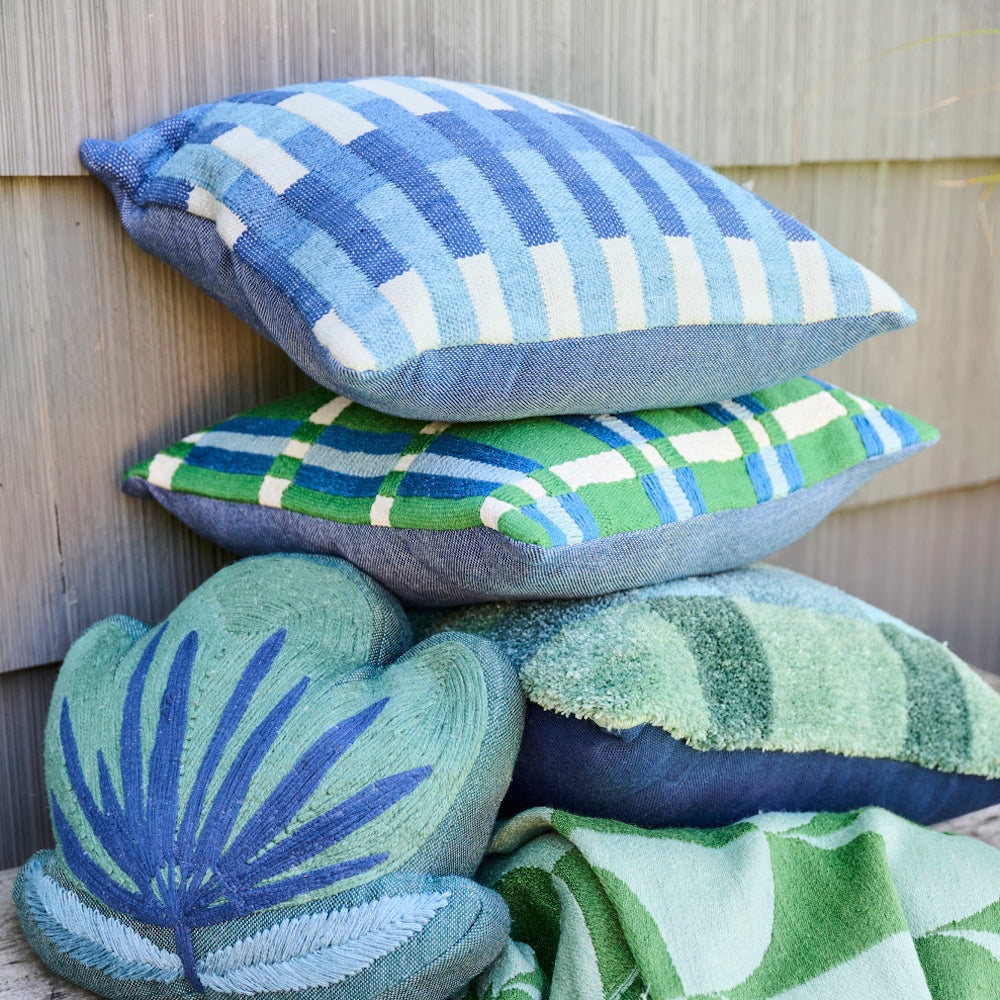 Stacked blue and green woven indoor-outdoor pillows with embroidery and leaf patterns on a rustic wooden bench.
