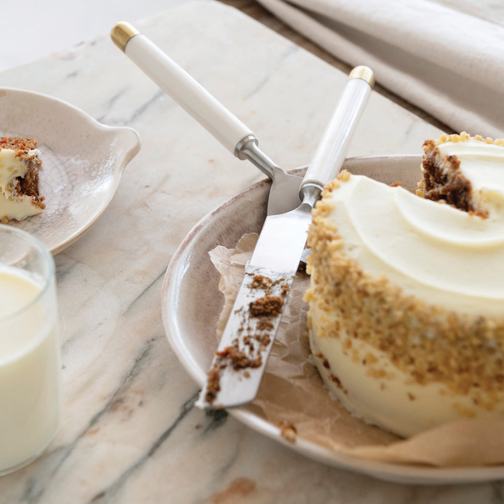 Lifestyle view of the stainless steel cake server and knife set with white and gold handles resting beside a frosted cake and a glass of milk.